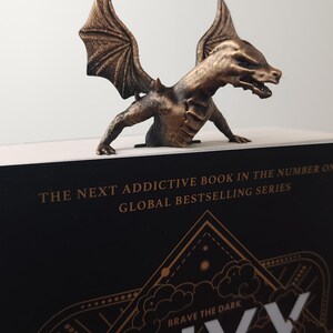 May include: Bronze dragon bookend perched on a book. The dragon has detailed wings and a menacing expression. The book has the title "ONYX STORM" and the text "THE NEXT ADDICTIVE BOOK IN THE NUMBER ONE GLOBAL BESTSELLING SERIES".