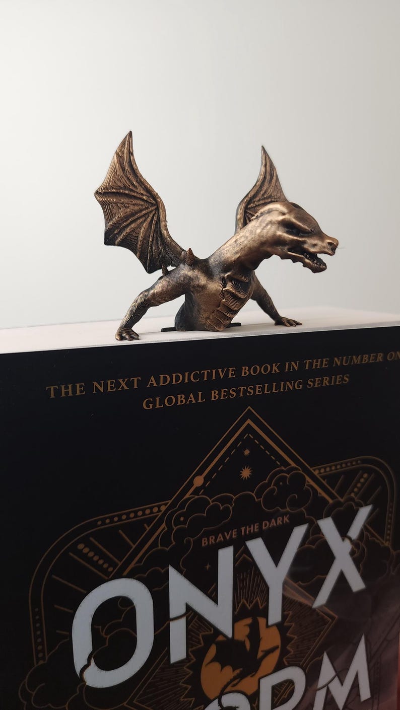 May include: Bronze dragon bookend perched on a book. The dragon has detailed wings and a menacing expression. The book has the title "ONYX STORM" and the text "THE NEXT ADDICTIVE BOOK IN THE NUMBER ONE GLOBAL BESTSELLING SERIES".