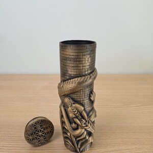 May include: A bronze-colored, cylindrical container with a detailed relief of a dragon wrapped around a tower. The container has a matching lid with a geometric pattern. The design is intricate and detailed.