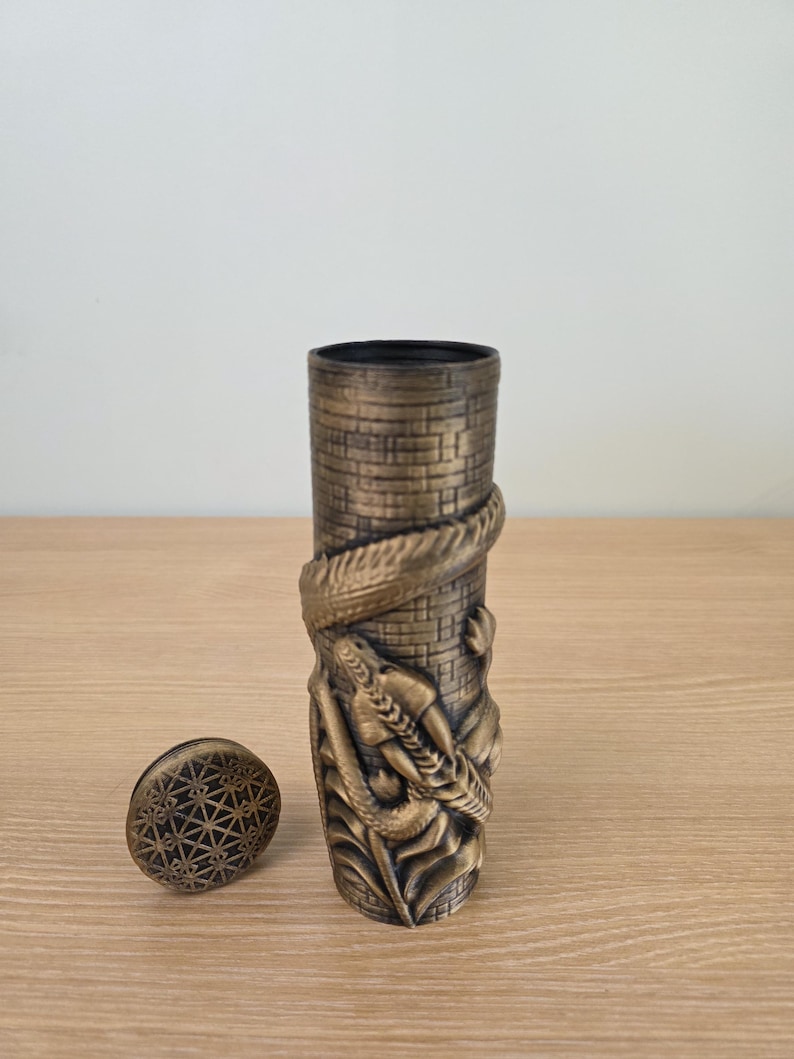 May include: A bronze-colored, cylindrical container with a detailed relief of a dragon wrapped around a tower. The container has a matching lid with a geometric pattern. The design is intricate and detailed.