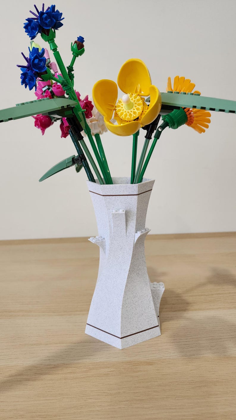 May include: A white ceramic vase with a brown rim and a textured surface holds a bouquet of colorful Lego flowers. The flowers include blue, pink, yellow, and green.