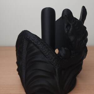 May include: Black dragon-shaped paper towel holder. The dragon is curled up with its wings spread out, and its head is tilted back. The holder is made of a textured plastic.