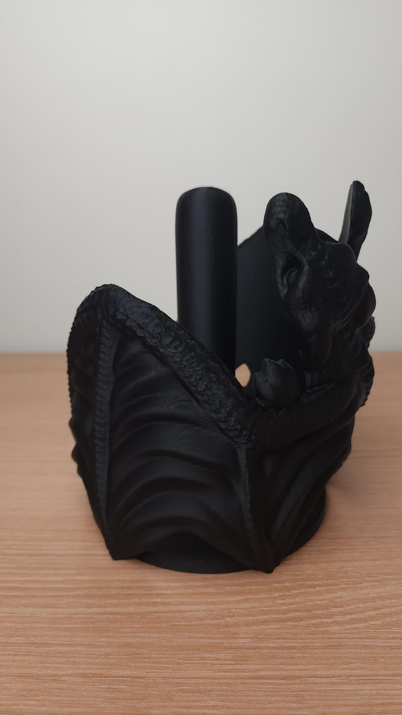 May include: Black dragon-shaped paper towel holder. The dragon is curled up with its wings spread out, and its head is tilted back. The holder is made of a textured plastic.