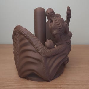 May include: A brown 3D printed dragon paper towel holder. The dragon is curled up with its wings spread out, and the paper towel roll sits inside the dragon's body.