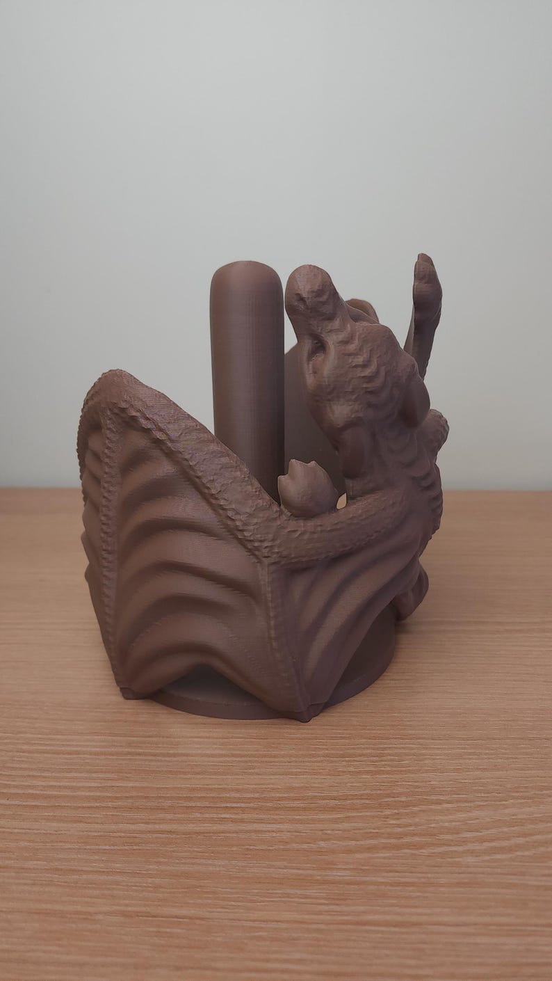 May include: A brown 3D printed dragon paper towel holder. The dragon is curled up with its wings spread out, and the paper towel roll sits inside the dragon's body.