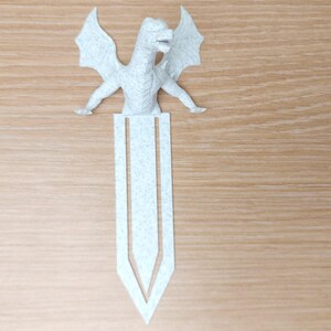 May include: A light gray dragon bookmark with outstretched wings and a detailed body. The bookmark is made of felt and has a pointed bottom. The dragon is positioned at the top of the bookmark, with its wings spread wide.