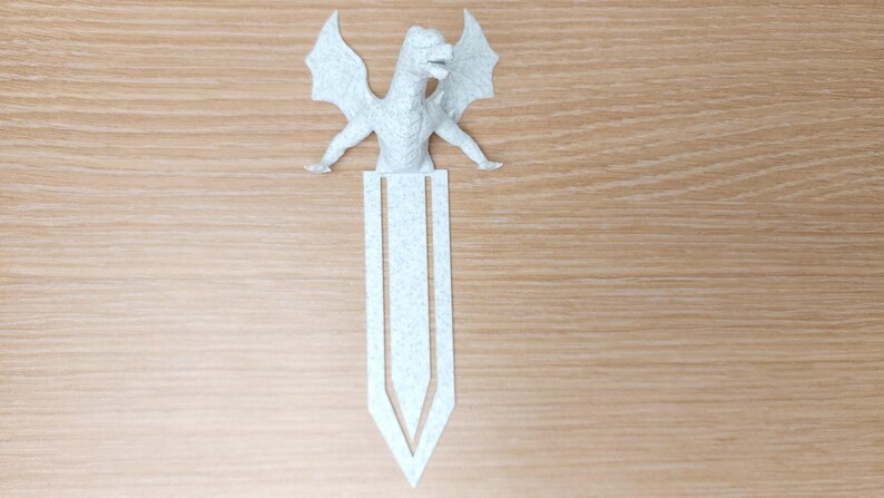 May include: A light gray dragon bookmark with outstretched wings and a detailed body. The bookmark is made of felt and has a pointed bottom. The dragon is positioned at the top of the bookmark, with its wings spread wide.