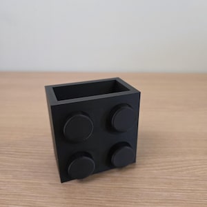 Peut inclure: Un r&eacute;cipient noir de forme carr&eacute;e ressemblant &agrave; une grosse brique de construction. Le r&eacute;cipient comporte quatre tenons circulaires sur un c&ocirc;t&eacute; et un int&eacute;rieur creux. L'objet est en mat&eacute;riau noir mat et repose sur une surface en bois.