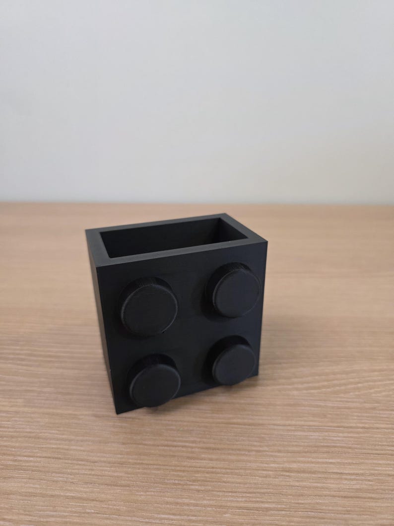 Peut inclure: Un r&eacute;cipient noir de forme carr&eacute;e ressemblant &agrave; une grosse brique de construction. Le r&eacute;cipient comporte quatre tenons circulaires sur un c&ocirc;t&eacute; et un int&eacute;rieur creux. L'objet est en mat&eacute;riau noir mat et repose sur une surface en bois.