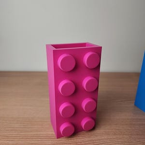 K&ouml;nnte beinhalten: Ein leuchtend rosa rechteckiger Beh&auml;lter, der einem gro&szlig;en Baustein &auml;hnelt. Der Beh&auml;lter hat acht kreisf&ouml;rmige Noppen auf einer Seite und einen hohlen Innenraum. Das Objekt besteht aus einem glatten Material und steht auf einer Holzoberfl&auml;che.