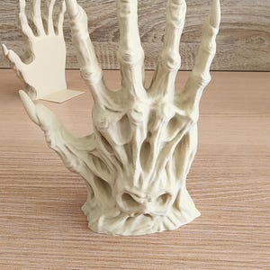 Peut inclure: Une sculpture d&eacute;taill&eacute;e, blanc cass&eacute;, d'une main aux doigts longs et pointus. La main a une apparence textur&eacute;e et squelettique, avec une structure osseuse visible et des espaces creux. Une deuxi&egrave;me sculpture de main plus petite est visible en arri&egrave;re-plan.