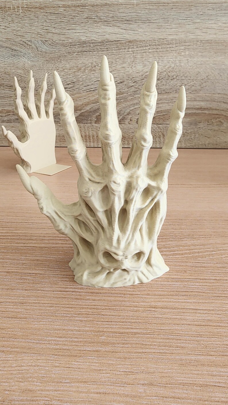 Peut inclure: Une sculpture d&eacute;taill&eacute;e, blanc cass&eacute;, d'une main aux doigts longs et pointus. La main a une apparence textur&eacute;e et squelettique, avec une structure osseuse visible et des espaces creux. Une deuxi&egrave;me sculpture de main plus petite est visible en arri&egrave;re-plan.