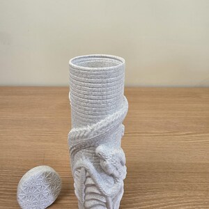 May include: A light gray cylindrical vase with a textured surface and a detailed snake wrapped around it. The vase has a matching oval lid with a geometric pattern. The design is a unique decorative piece.