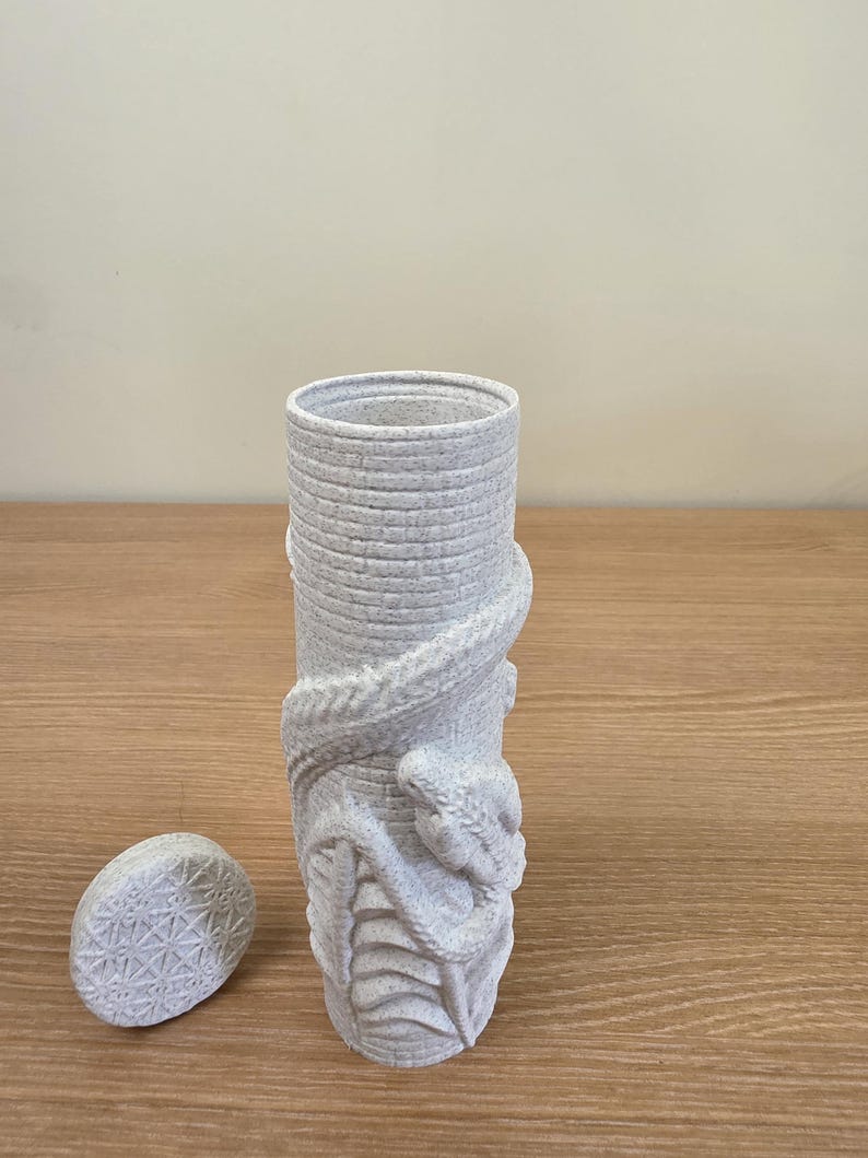 May include: A light gray cylindrical vase with a textured surface and a detailed snake wrapped around it. The vase has a matching oval lid with a geometric pattern. The design is a unique decorative piece.