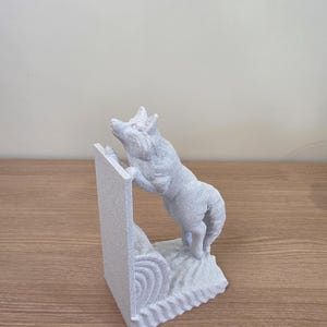 Op de afbeelding: Een lichtgrijze sculptuur van een wolf die een getextureerde muur beklimt. De wolf staat op zijn achterpoten en reikt omhoog. De sculptuur is gemaakt van een gespikkeld materiaal en staat op een houten oppervlak.