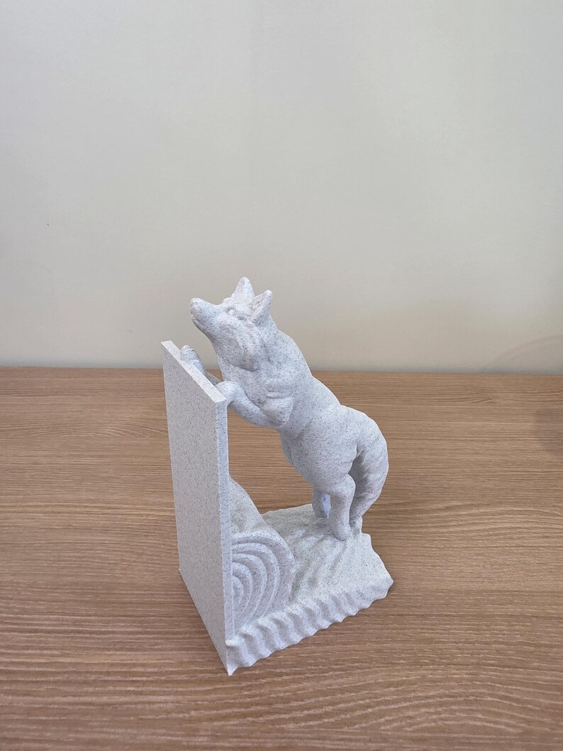 Op de afbeelding: Een lichtgrijze sculptuur van een wolf die een getextureerde muur beklimt. De wolf staat op zijn achterpoten en reikt omhoog. De sculptuur is gemaakt van een gespikkeld materiaal en staat op een houten oppervlak.