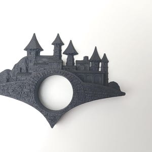 May include: A black 3D printed bookmark shaped like a castle with a round hole in the center. The castle is on a hill with a bridge.