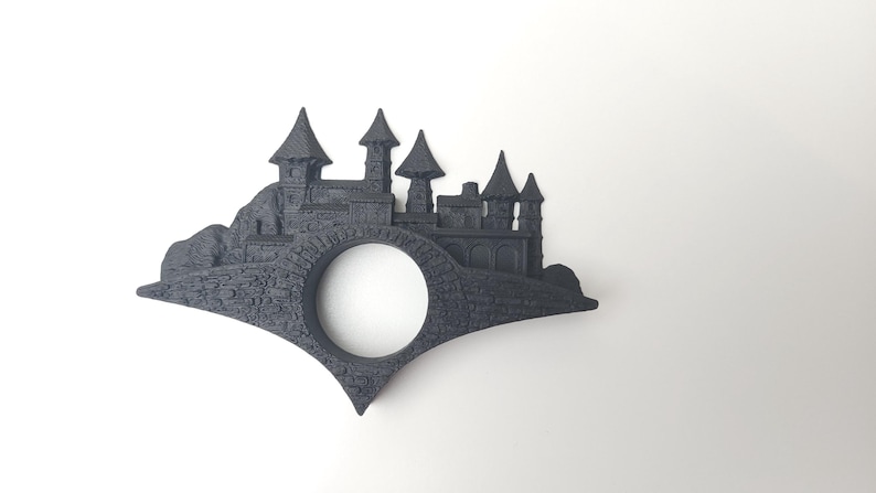 May include: A black 3D printed bookmark shaped like a castle with a round hole in the center. The castle is on a hill with a bridge.