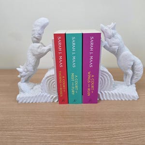 Op de afbeelding: Witte decoratieve boekensteunen met vosachtige figuren die drie boeken flankeren. De boeken hebben rode, turquoise en paarse ruggen met de titels "A Court of Thorns and Roses", "A Court of Mist and Fury" en "A Court of Wings and Ruin".
