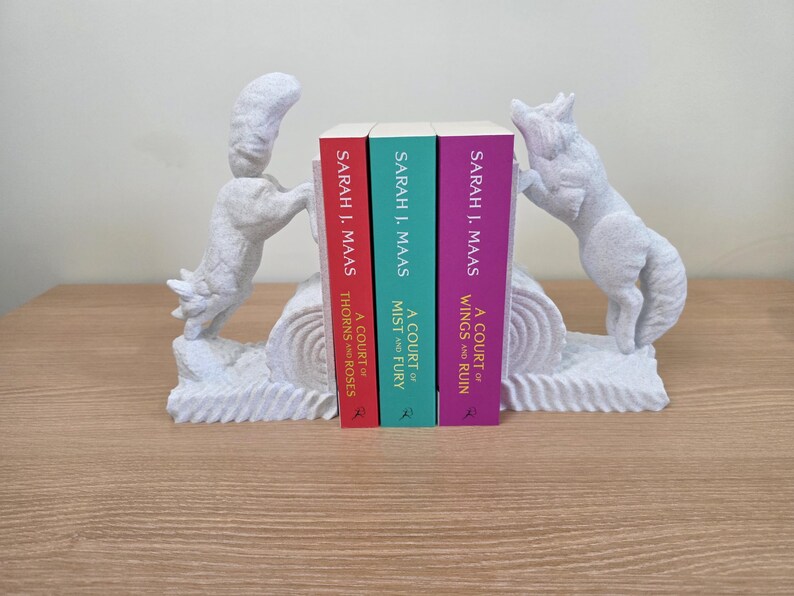Op de afbeelding: Witte decoratieve boekensteunen met vosachtige figuren die drie boeken flankeren. De boeken hebben rode, turquoise en paarse ruggen met de titels "A Court of Thorns and Roses", "A Court of Mist and Fury" en "A Court of Wings and Ruin".