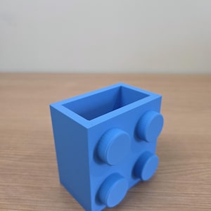 Peut inclure: Un r&eacute;cipient rectangulaire bleu clair avec quatre plots circulaires en relief sur un c&ocirc;t&eacute;, ressemblant &agrave; une grosse brique de construction. Le dessus ouvert r&eacute;v&egrave;le un int&eacute;rieur vide. L'objet est en mat&eacute;riau mat.