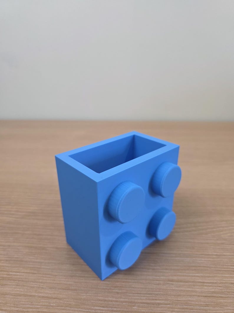 Peut inclure: Un r&eacute;cipient rectangulaire bleu clair avec quatre plots circulaires en relief sur un c&ocirc;t&eacute;, ressemblant &agrave; une grosse brique de construction. Le dessus ouvert r&eacute;v&egrave;le un int&eacute;rieur vide. L'objet est en mat&eacute;riau mat.