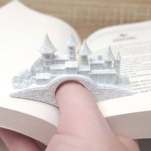 May include: White 3D printed castle bookmark with a bridge that fits over a finger. The bookmark is sitting on a book.