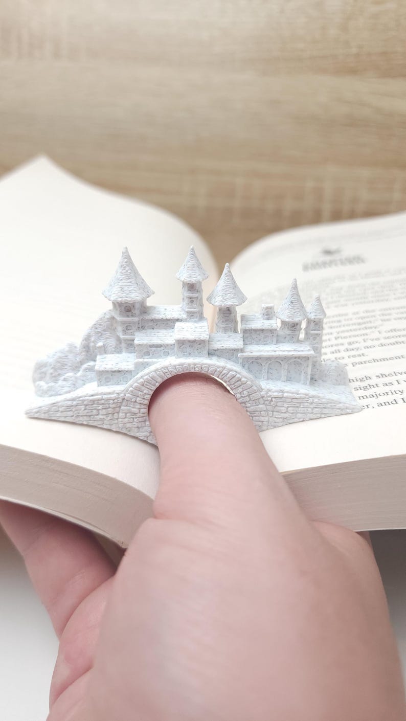 May include: White 3D printed castle bookmark with a bridge that fits over a finger. The bookmark is sitting on a book.