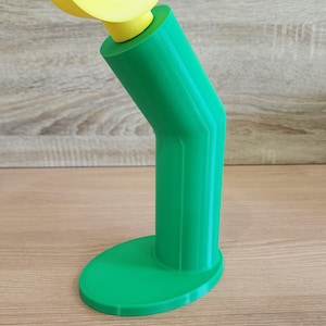 May include: A green 3D printed phone stand with a yellow plastic hook on top. The stand has a rounded base and a curved neck.