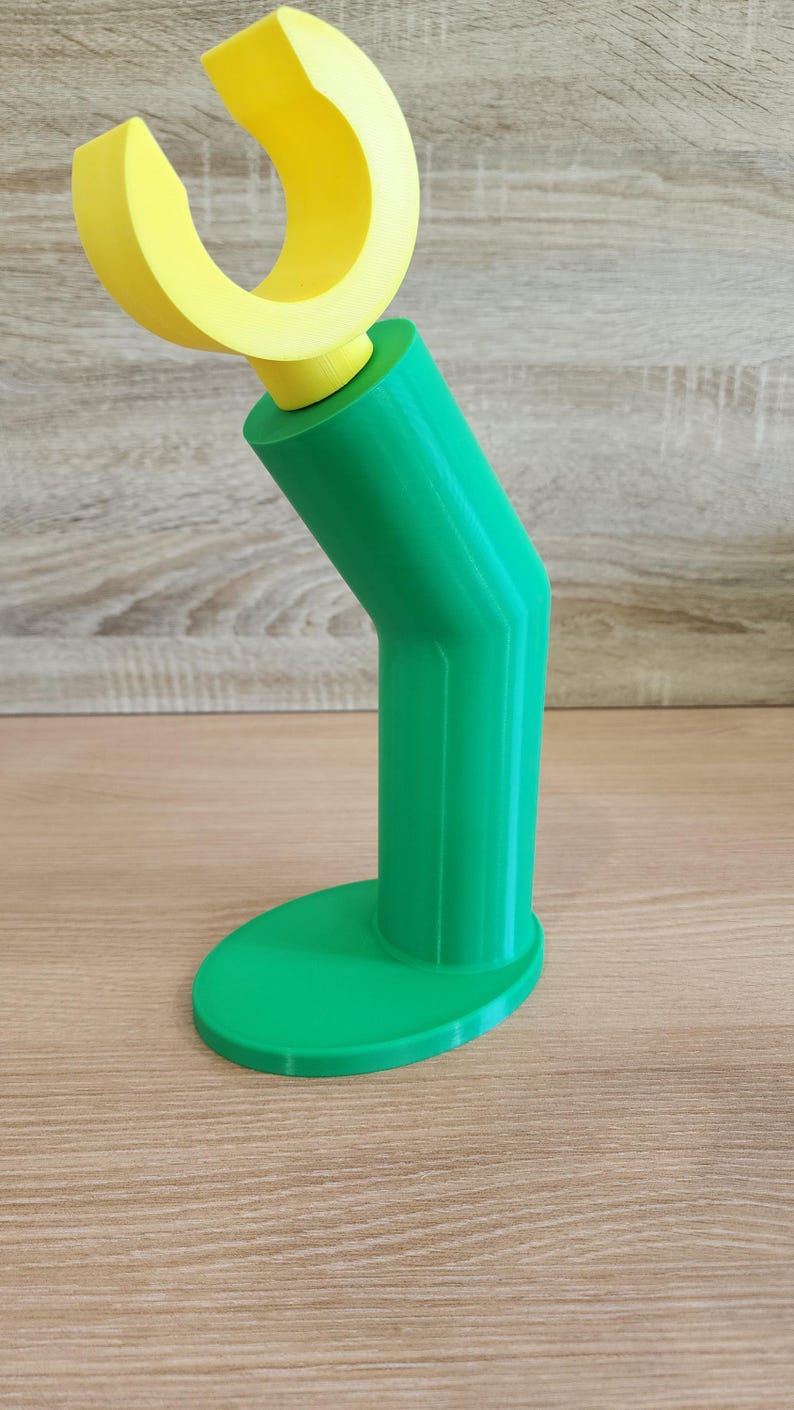 May include: A green 3D printed phone stand with a yellow plastic hook on top. The stand has a rounded base and a curved neck.
