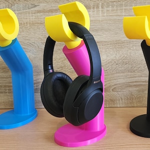 May include: Three headphone stands in different colors: blue, pink, and black. Each stand has a yellow plastic hook on top to hold the headphones. The pink stand has a pair of black headphones on it.