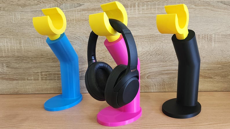 May include: Three headphone stands in different colors: blue, pink, and black. Each stand has a yellow plastic hook on top to hold the headphones. The pink stand has a pair of black headphones on it.