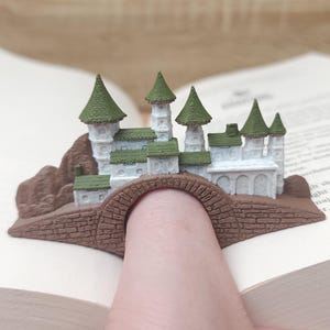 May include: A brown and green 3D printed castle bookmark with a bridge that fits over a finger. The castle has white walls and green roofs.