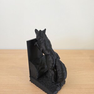 Op de afbeelding: Een zwarte vossen boekensteun sculptuur. De vos wordt afgebeeld in een klimhouding, met zijn voorpoten op een rechthoekige steun. De basis van de boekensteun heeft een getextureerd, grasachtig uiterlijk. De sculptuur is gemaakt van een effen zwart materiaal.