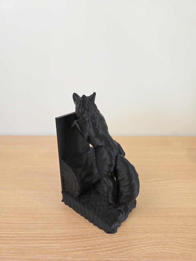 Op de afbeelding: Een zwarte vossen boekensteun sculptuur. De vos wordt afgebeeld in een klimhouding, met zijn voorpoten op een rechthoekige steun. De basis van de boekensteun heeft een getextureerd, grasachtig uiterlijk. De sculptuur is gemaakt van een effen zwart materiaal.