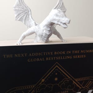 May include: A white dragon figurine with detailed wings and a textured surface, perched atop a book. The book has the title "ONYX" and the text "Brave the Dark" in gold lettering. The overall theme is fantasy and literature.