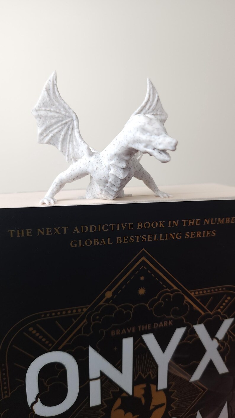 May include: A white dragon figurine with detailed wings and a textured surface, perched atop a book. The book has the title "ONYX" and the text "Brave the Dark" in gold lettering. The overall theme is fantasy and literature.