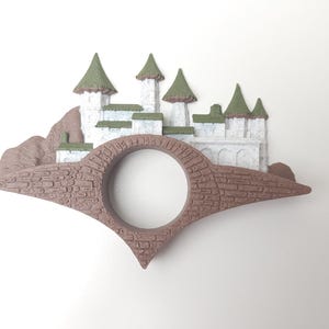 May include: A brown and white castle figurine with green roofs, perched on a brown bridge with a round opening in the center.