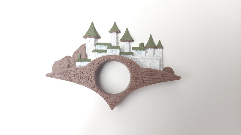 May include: A brown and white castle figurine with green roofs, perched on a brown bridge with a round opening in the center.