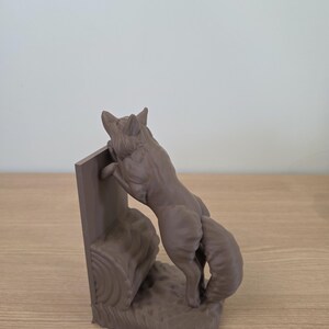 Op de afbeelding: Een bruine vossensculptuur, staand op zijn achterpoten, lijkt over een boomstam te klimmen. De vos maakt deel uit van een boekensteunset, met een rechthoekige steun. De sculptuur is gemaakt van een mat materiaal.