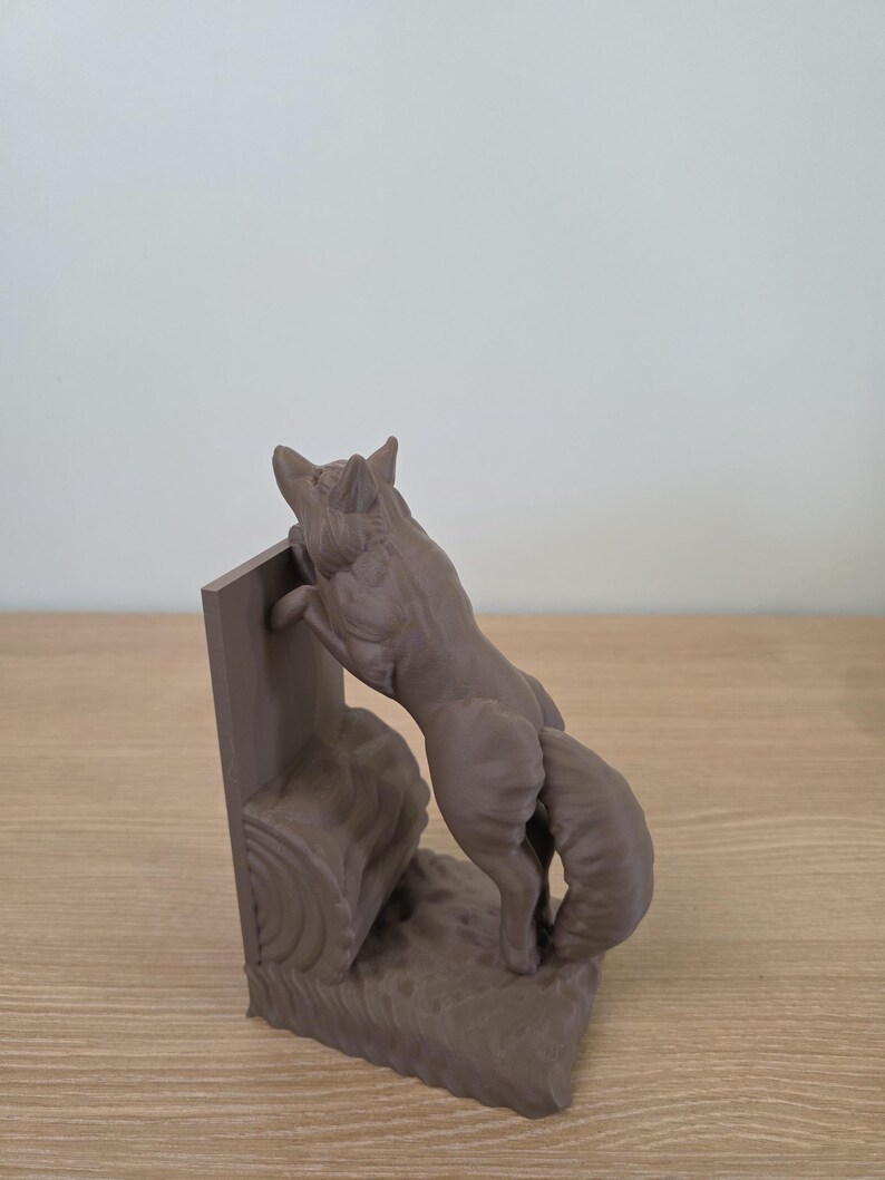 Op de afbeelding: Een bruine vossensculptuur, staand op zijn achterpoten, lijkt over een boomstam te klimmen. De vos maakt deel uit van een boekensteunset, met een rechthoekige steun. De sculptuur is gemaakt van een mat materiaal.