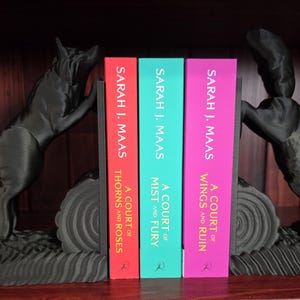 Op de afbeelding: Zwarte wolf-boekensteunen flankeren drie boeken met kleurrijke ruggen. De boeken heten "A Court of Thorns and Roses", "A Court of Mist and Fury" en "A Court of Wings and Ruin" van Sarah J. Maas.