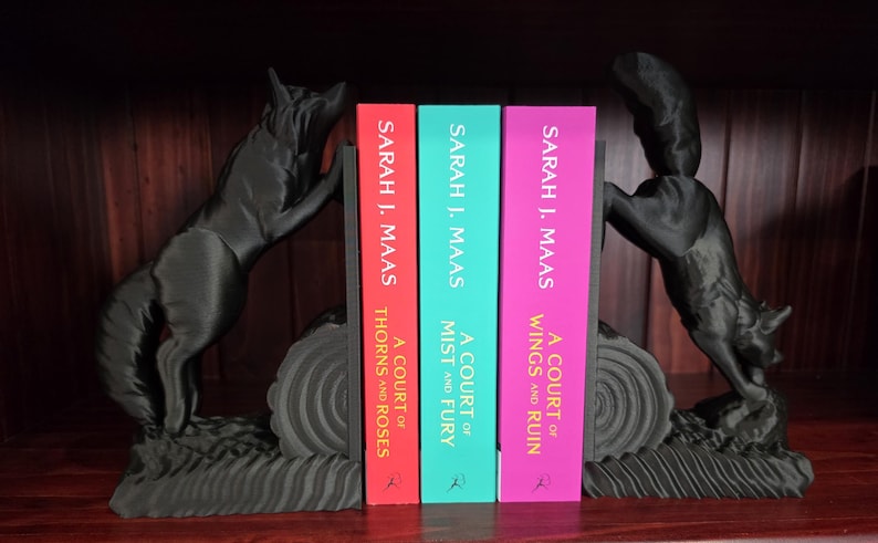 Op de afbeelding: Zwarte wolf-boekensteunen flankeren drie boeken met kleurrijke ruggen. De boeken heten "A Court of Thorns and Roses", "A Court of Mist and Fury" en "A Court of Wings and Ruin" van Sarah J. Maas.