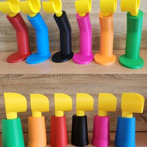 May include: Two rows of 3D printed phone stands in various colors, including red, blue, black, pink, orange, and green. The stands have a yellow hook at the top and a base in a variety of shapes.