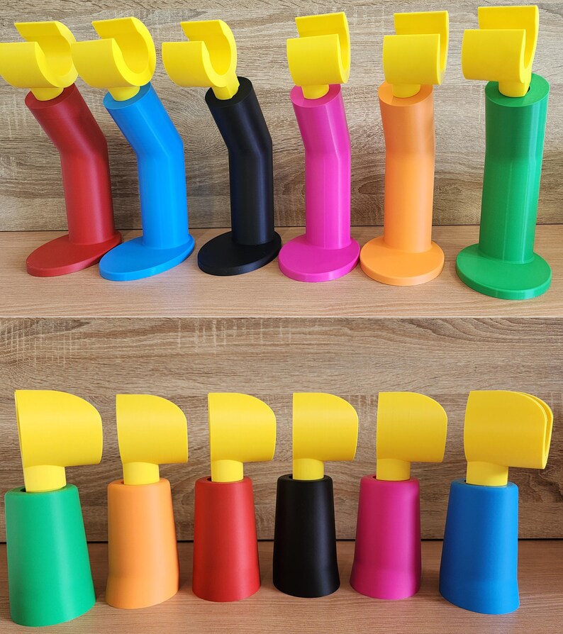 May include: Two rows of 3D printed phone stands in various colors, including red, blue, black, pink, orange, and green. The stands have a yellow hook at the top and a base in a variety of shapes.