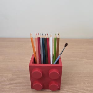Peut inclure: Un porte-crayons rouge carr&eacute;, con&ccedil;u pour ressembler &agrave; une brique de construction, rempli de crayons de couleur et d'un petit pinceau. Le support comporte quatre plots circulaires en relief &agrave; l'avant et repose sur une surface en bois.