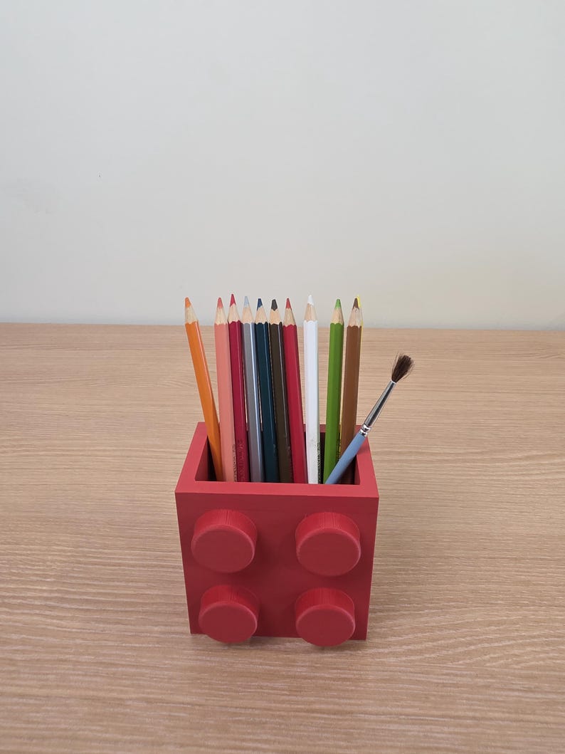 Peut inclure: Un porte-crayons rouge carr&eacute;, con&ccedil;u pour ressembler &agrave; une brique de construction, rempli de crayons de couleur et d'un petit pinceau. Le support comporte quatre plots circulaires en relief &agrave; l'avant et repose sur une surface en bois.