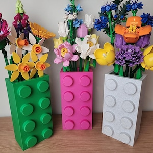 May include: Three colourful vases, resembling large building blocks, hold vibrant, artificial flowers. The vases are green, pink, and grey, and the flowers include pink, yellow, and purple blooms. The vases are approximately 25 cm tall.