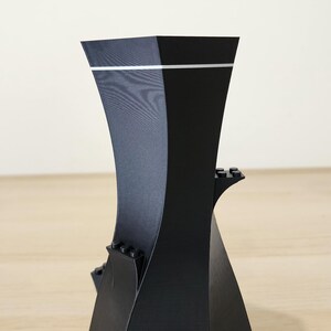 May include: Black 3D printed phone stand with a geometric design and white accents. The stand has multiple slots for holding phones and other devices.