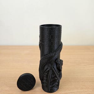 May include: A black cylindrical vase with a detailed dragon design wrapped around it. The vase has a matching lid with a geometric pattern. The vase is approximately 6 inches tall.